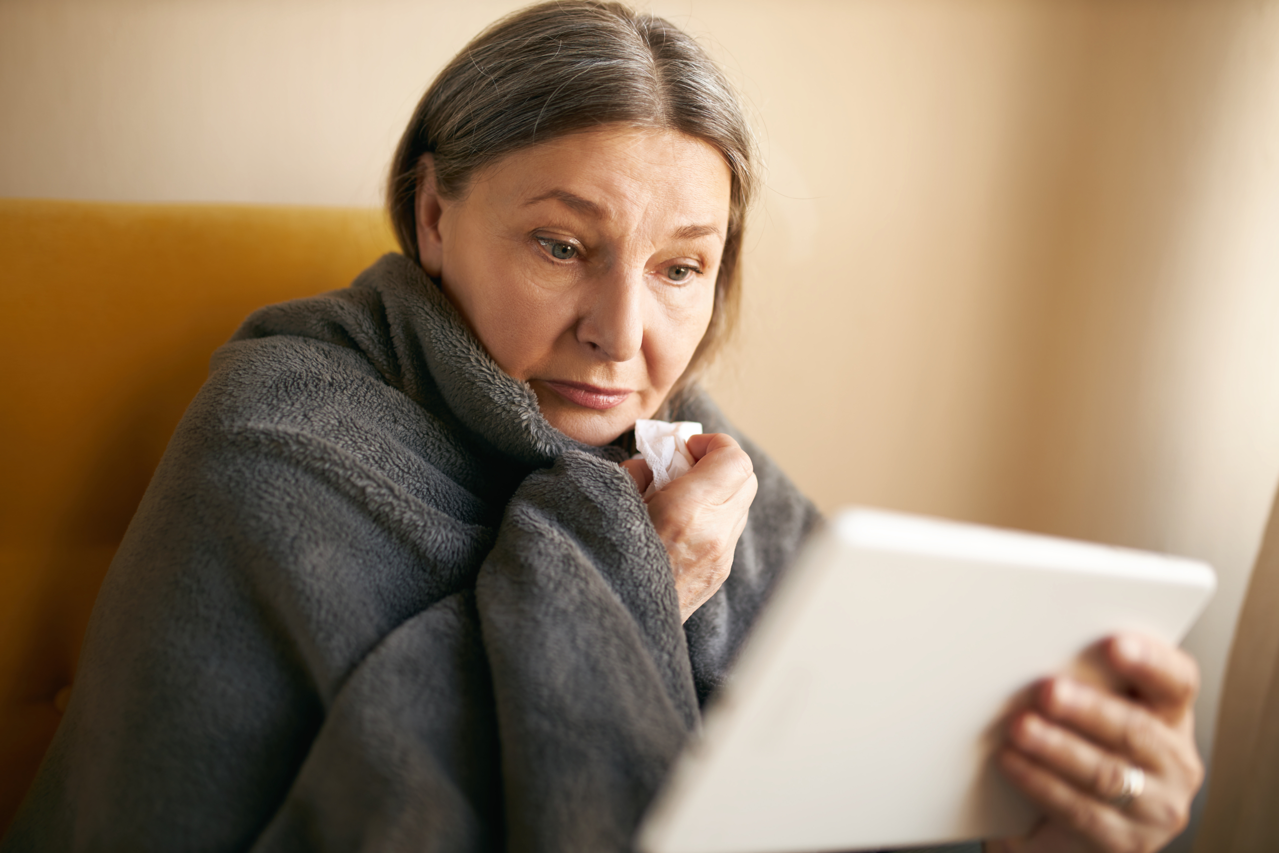 Sick gray haired retired woman with running nose sitting at home wrapped in plaid holding digital tablet having online consultation with doctor using video conferencing platform application